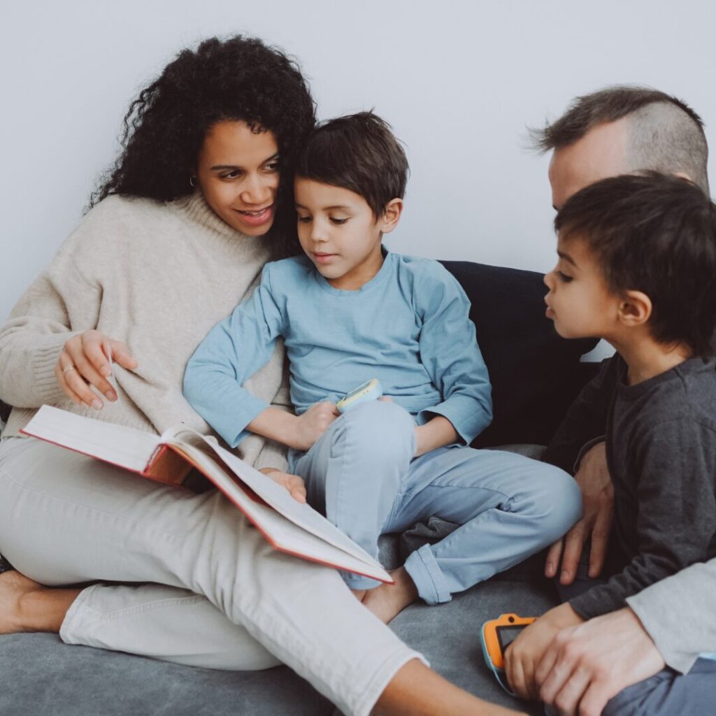A family sits together, one child in mom's lap and another sitting with dad to look at the book mom is reading.