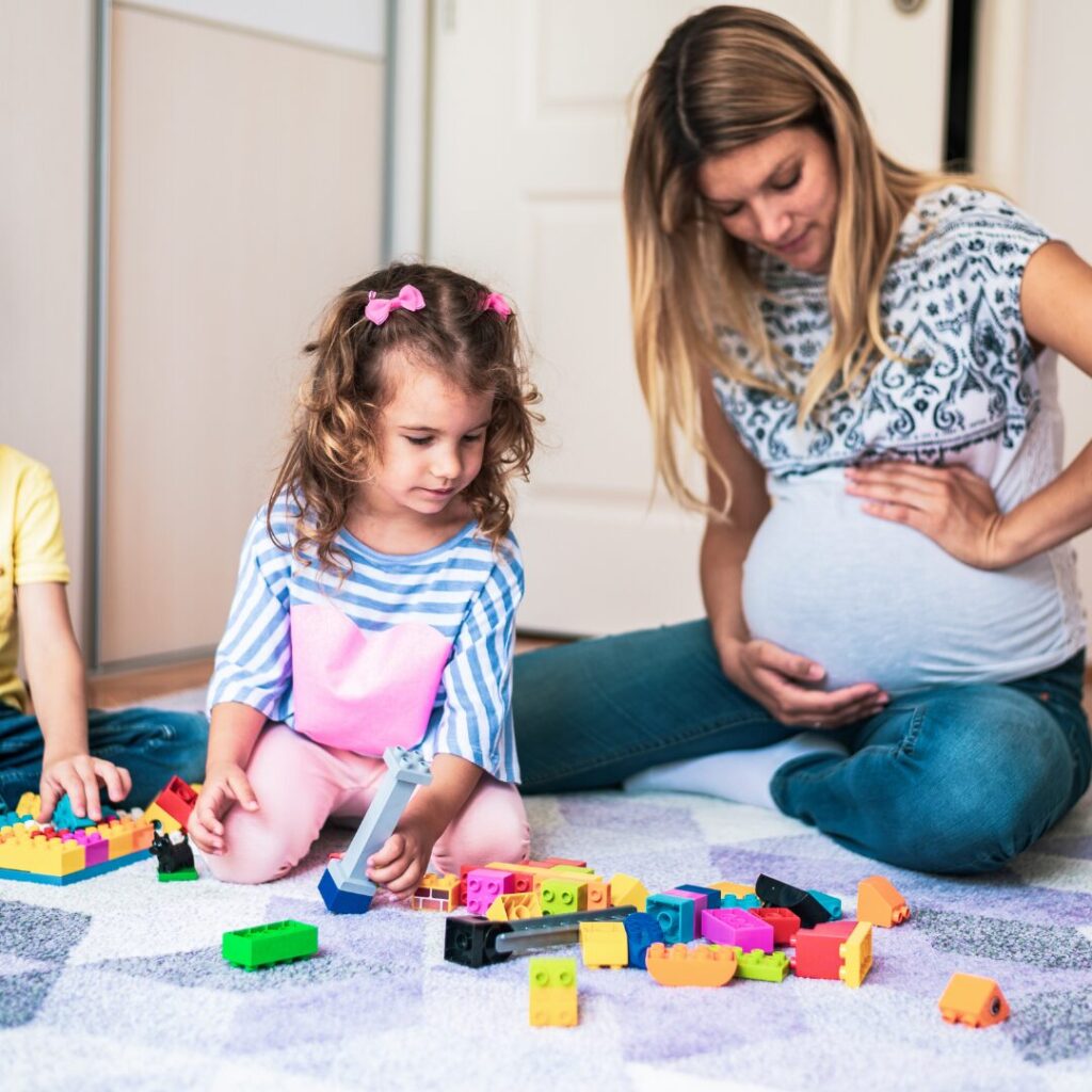 A pregnant mom holds her belly while playing on the floor with her two older children