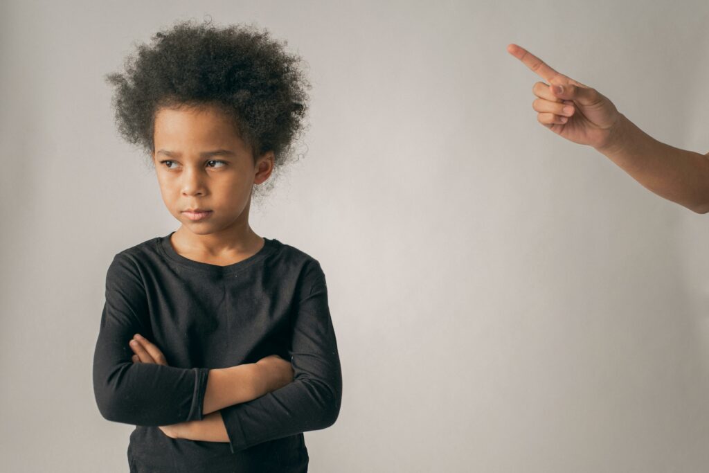 pexels-gabby-k-7114747 A child frowning and looking away, arms crossed, while an adult hand comes in from the side of the frame, finger pointed