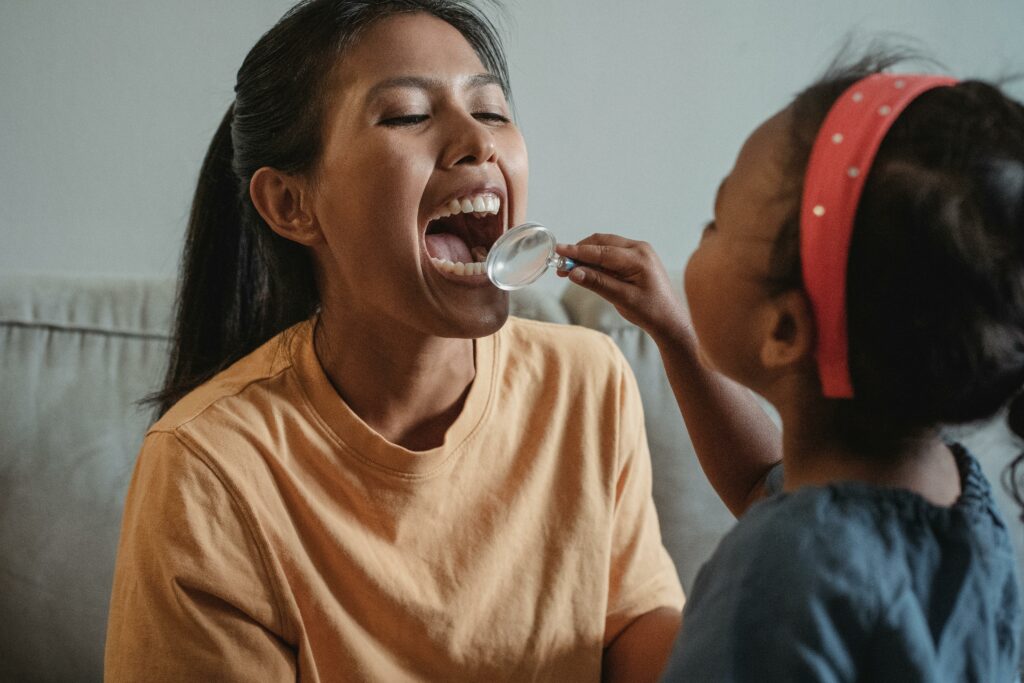 A child uses a magnifying glass to look inside her mother's open, smiling mouth.