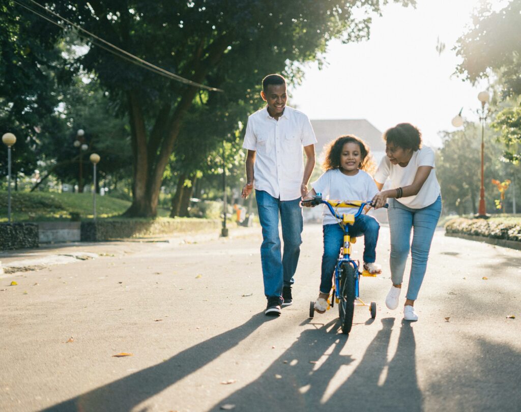 A family is outside in the sunlight. A mother leans over a bicycle with training wheels, teaching her daughter how to ride, while dad follows behind laughing proudly.