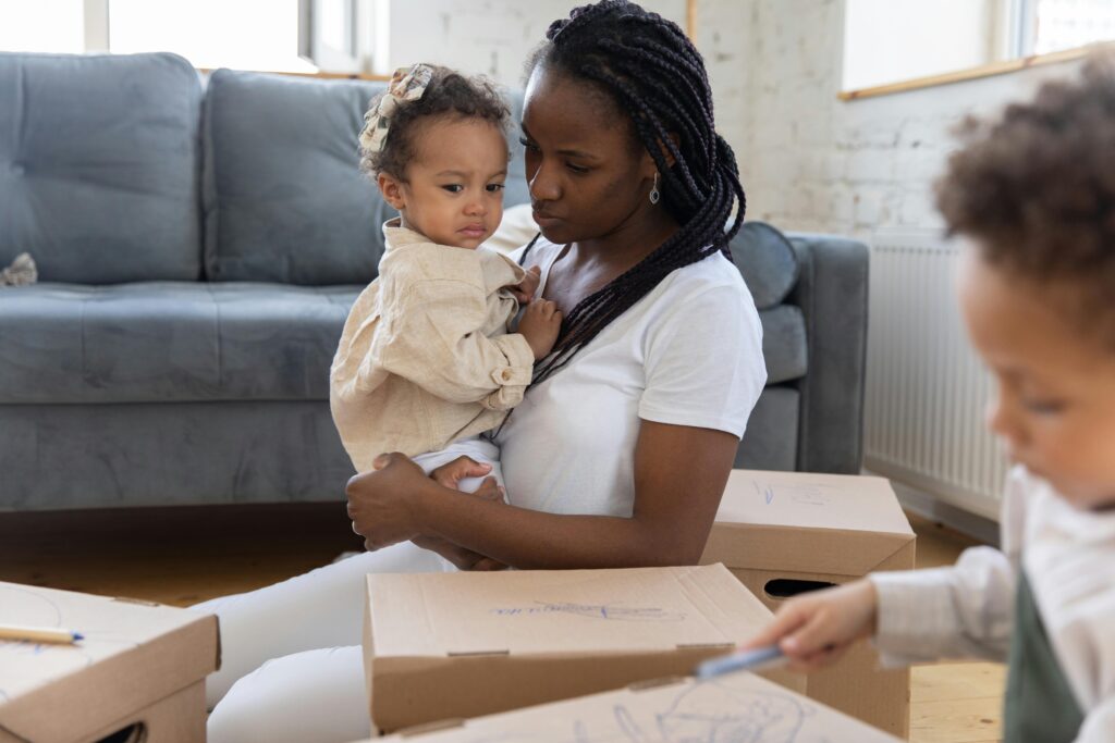 pexels-ekaterina-bolovtsova-4868630 A mother sits with her child in her arms looking lost, while her older toddler colours on the lids of cardboard boxes