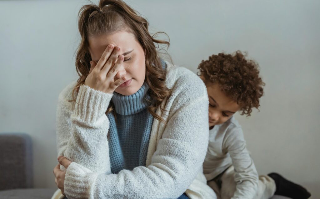A mother sitting with her face in her hand, her child snuggled against her back