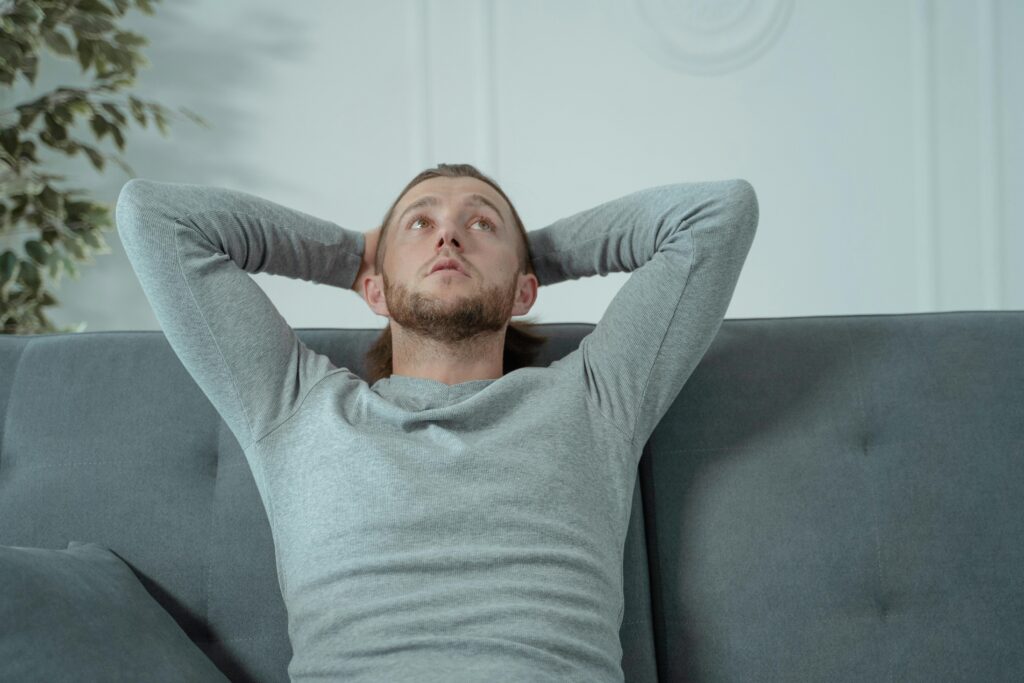 A father sits on a couch, his head behind his hands, staring at the ceiling looking stressed