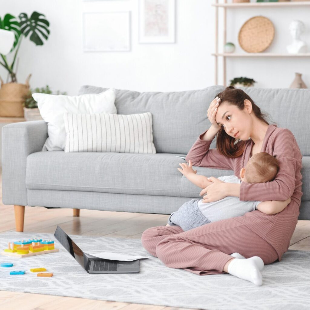 A mom sitting with her toddler across her lap, laptop open on the floor before her, looking tired.