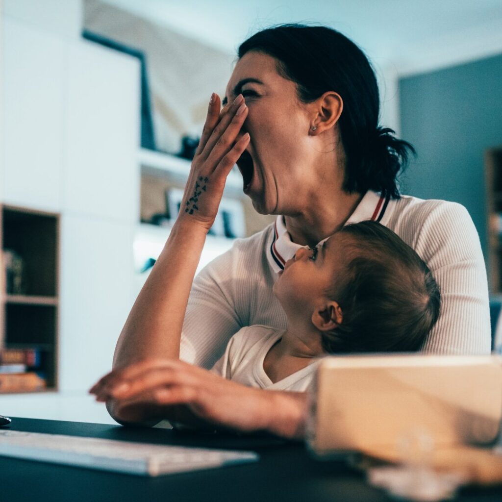 A mom yawns hugely while her baby looks up at her from her lap, while she's sitting at a home office desk doing some work on her laptop