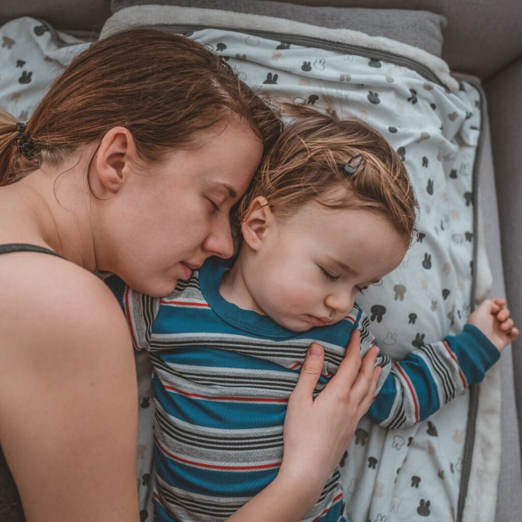 A tired mom has fallen asleep next to her sleeping child on a soft bed.