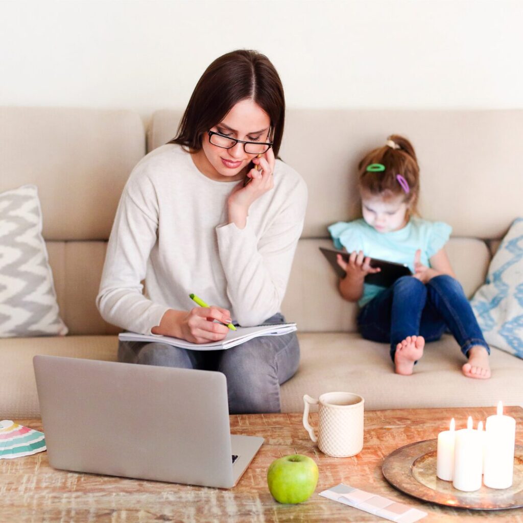 A mother writes notes on a notebook, a laptop before her, while she is on the phone; behind her, her young daughter is sitting on a couch, playing on a tablet.