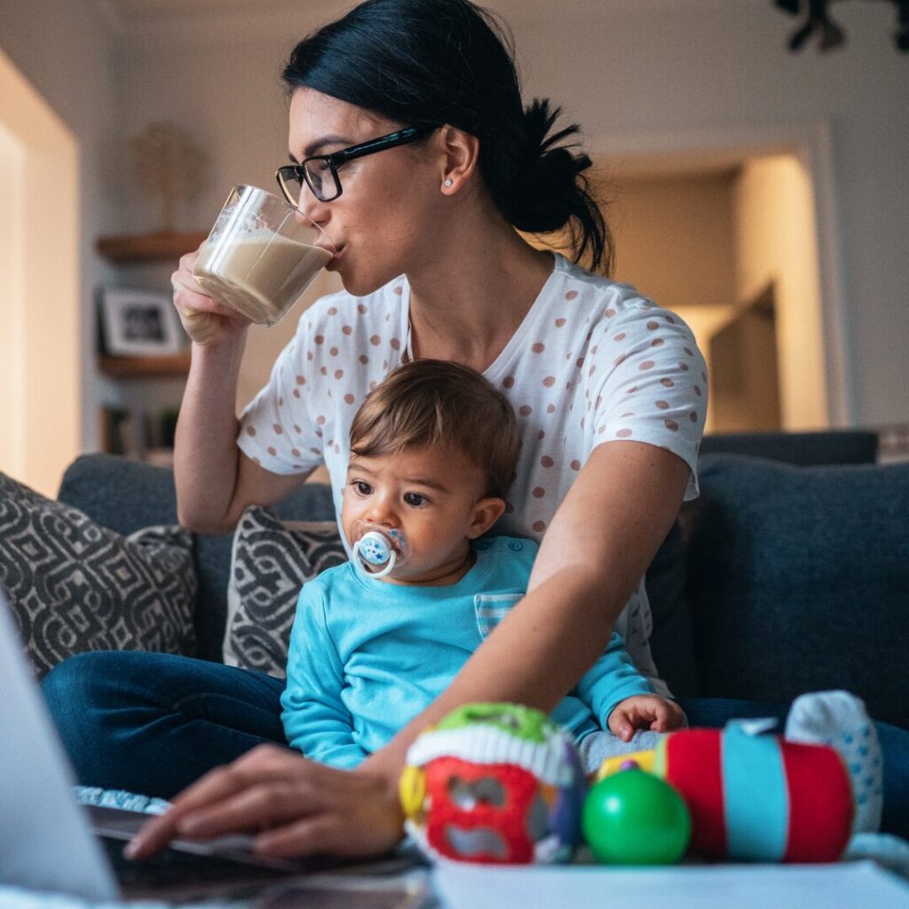 A mother sipping a mug of tea with her toddler in her lap while she sits cross-legged, typing one-handed on the laptop before her.