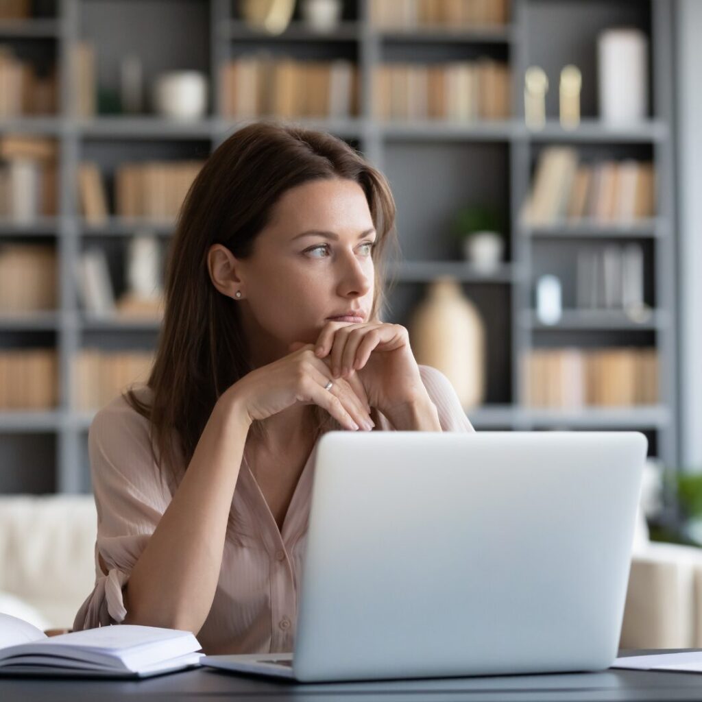 A mom is at work in her office alone, sitting at her laptop, and yet looking out the nearby window, thinking about what she'll need to do that evening at home when she returns to mom-life.